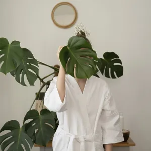 Eine Frau in einem weißen Bademantel hält ein großes Monstera-Blatt vor ihr Gesicht. Eine Frau in einem weißen Bademantel hält ein großes Monstera-Blatt vor ihr Gesicht.