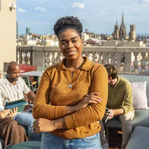 Young black woman with an afro hairstyle smiles at the camera on a modern rooftop terrace while diverse colleagues work