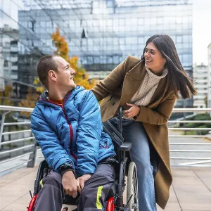 Young man in a wheelchair smiling and chatting with a supportive woman on a sunny urban street