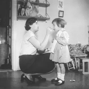 A young mother administers medicine to her daughter with a spoon in her mouth in 1951