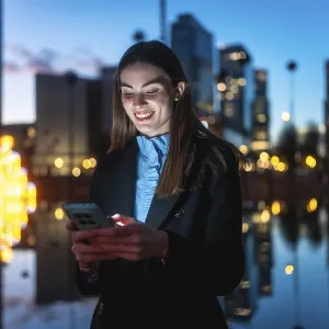Young professional woman smiling happily while interacting with her mobile phone against a blurred city background with Young professional woman smiling happily while interacting with her mobile phone against a blurred city background with