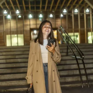 Young woman in a trench coat smiling and looking at her smartphone while standing on modern urban stairs at night Young woman in a trench coat smiling and looking at her smartphone while standing on modern urban stairs at night