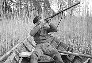 In the 1930s. A man sits in a rowing boat and aims his rifle upwards to shoot ducks