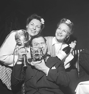 1940s photographer. A man with a Rolleiflex camera and flash aims at something with two girls haning on his shoulders