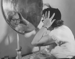 In the 1950s. A lady looks her self in the wall mirror to see that her hat looks good on her