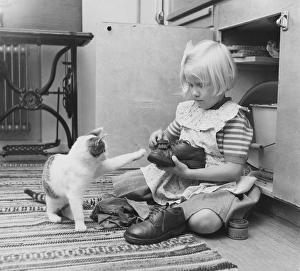 In the 1950s. A little girl is sitting on the kitchen floor polishing her shoes and a cat stretches out it's paw