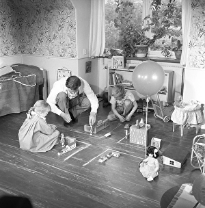 In the 1950s. A man with two children are playing together on the floor with cars and houses
