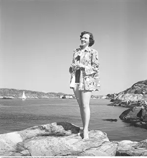 1950s summer fashion. A young woman in a bikini with a light short coat pictured standing on a cliff on the coast