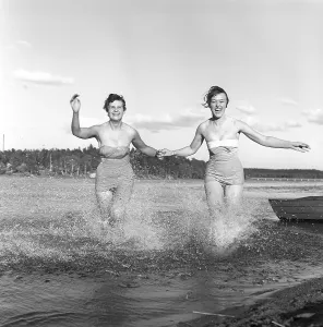 1950s summer. Two young women runs on the shallow part of the beach and the water splashes