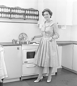 In the 1950s. A woman in a typical 1950s kitchen with wooden cabinets and a set of spices on shelfes above the cooker
