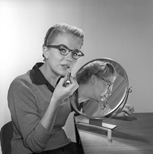 In the 1950s. A young woman with a mirror putting lipstick on. She has the glasses and bows typical of the 1950s. 1958. Kristoffersson ref CB99-12