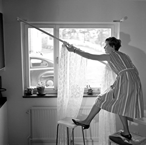 1960s home decorating. A woman at home putting up the curtains in the room window. All by herself she balances on a chair and a stool to manage it. Sweden 1966