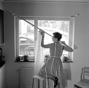 1960s home decorating. A woman at home putting up the curtains in the room window. All by herself she balances on a chair and a stool to manage it. Sweden 1966