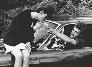 In the 1960s. A women is sitting on the hood of a car and a man reaches out with a cigarette lighter to light hers