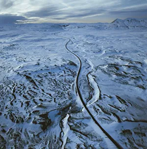 Aerial view of a long road winding through a vast snowy landscape with distant mountains under a cloudy sky