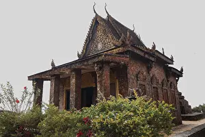An ancient red brick temple with ornate carvings surrounded by lush greenery and flowers under a cloudy sky