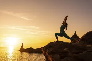 Anjaneyasana, a woman doing meditation and yoga exercises on a rock at sunset next to a lighthouse in the sea