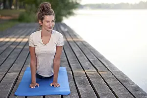 Attractive young woman working out on mat on a wooden deck above a lake or river doing a yoga pose smiling