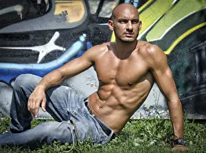 Bald young man shirtless outdoors sitting in front of graffiti wall on grass