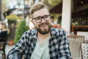 Bearded man in glasses laughing joyfully while sitting at a cafe table outdoors