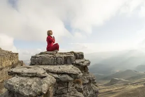 A beautiful girl meditates in a lotus pose sitting on a rock above the clouds against the backdrop of the setting sun