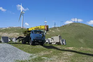 A blue crane lorry stands on a green construction site next to a wind turbine on a hill