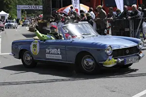 A blue open-top vintage rally car with a smiling driver in front of spectators