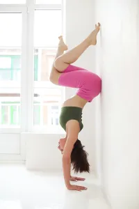 Full body of focused slim female female performing handstand near wall while practicing yoga in light room by