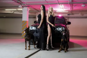 Full body side view of young female in black dress standing with black Doberman and looking at camera