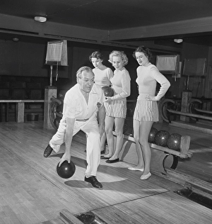Bowling in the 1950s. A man is throwing the bowling ball and three young women are standing beside and looking. they are dressed in short striped skirts and jumpers