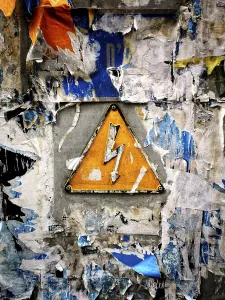 A bright yellow warning sign featuring a lightning bolt is mounted on a wall heavily covered with faded