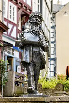 Framed Print : Bronze sculpture of a man with hat holding a card in a historic old town with half-timbered houses Framed Print : Bronze sculpture of a man with hat holding a card in a historic old town with half-timbered houses