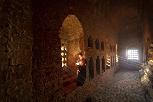Category Mountain Landscapes Prints: Buddhist young monk in red robe reading standing in front of rays of light in a temple