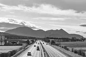 Busy motorway in front of a mountain backdrop in black and white with cloudy sky