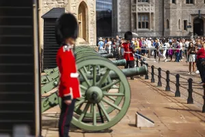 Ceremonial guards in red uniforms stand with cannons as tourists observe a historic site at The Tower of London