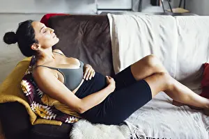Cheerful young brunette latin pregnant sportswoman sitting on couch in light room in relaxing pose doing her yoga
