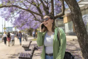 A cheerful young woman with sunglasses and a green jacket talks on her phone during a sunny day on the riverfront