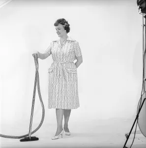 Cleaning day in the 1960s. A woman is standing holding a vacuum cleaner