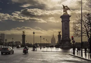 Clouds gather above the iconic Alexandre III Bridge as vehicles and people navigate the bustling streets of Paris