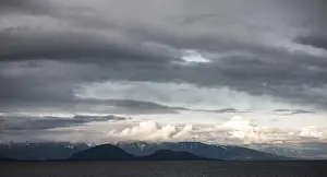 Clouds hover over distant mountains and sea in a moody landscape as seen from a passenger ferry