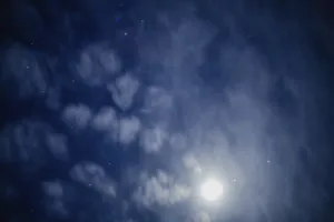 A cloudy night sky with shining stars and a bright moon partially hidden by clouds