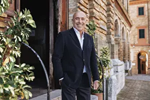 A confident businessman in a black blazer smiles while standing outside a historic brick building