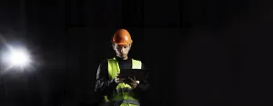 A construction worker in safety gear uses a tablet under spotlight against a dark backdrop