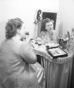 Cosmetics in the 1940s. A young woman at home is sitting at a make up desk and looking at herself in the mirror while putting on lipstick. There are many different kinds of cosmectics lying on the table in front of her. Sweden 1947