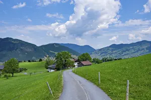 Framed Print : Country road leads through green meadows into a rural mountain landscape under a blue sky with white clouds