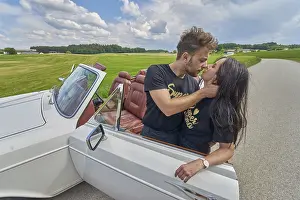 Couple kissing by a convertible car amidst a rustic countryside backdrop