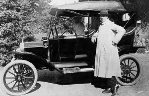 Couple, woman sits at the wheel of a vintage car and the man stands in front of the car