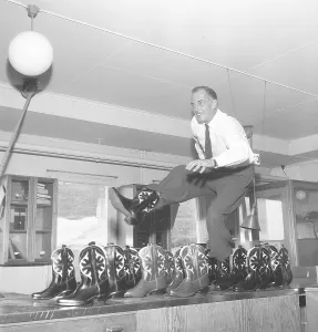 Cowboy boots. A man is playing around and dancing on a table with cowboy boots. Another dozen of cowboy boots are standing on the side. Sweden 1956. Photo Kristoffersson Ref BX20-6