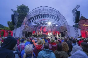 Crowd gathers in front of an open-air stage under the evening sky with light spectacle