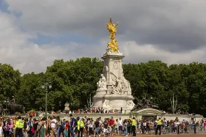 Crowd surrounds Victoria Memorial's golden statue amidst overcast skies and green trees in the City of Westminster